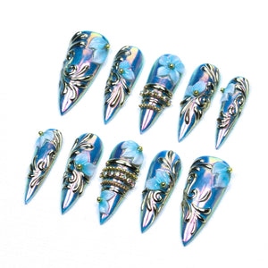A set of ten stiletto-shaped press-on nails featuring an iridescent blue finish. Each nail is decorated with raised gold filigree patterns, small light blue floral accents, and some include bands of embedded clear rhinestones.