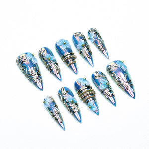 A set of ten stiletto-shaped press-on nails featuring an iridescent blue finish, silver filigree scrollwork, raised light blue floral accents, and small gold beads. Some nails include horizontal bands of clear rhinestones.
