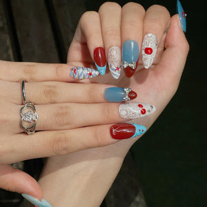 Almond-shaped nails featuring a festive design with red, blue, and white colors, intricate white lace patterns, small red star accents, and silver bow charms. A silver-toned ring with a heart-shaped cutout design is worn on the finger.