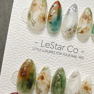 A set of almond-shaped press-on nails featuring floral patterns, green and brown watercolor washes, and silver foil accents. The nails are displayed on a white textured card with the text LeStar Co -
