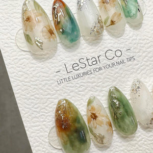 A set of almond-shaped press-on nails featuring floral designs, green and orange watercolor-style swirls, and gold foil accents. The nails are displayed on a white card with the text LeStar Co - LITTL