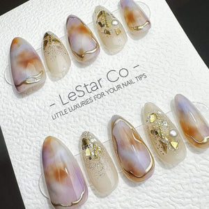 A set of ten almond-shaped press-on nails featuring a marbled design in shades of purple, brown, and cream, accented with gold foil flakes, glitter, and small white pearls. The nails are displayed on