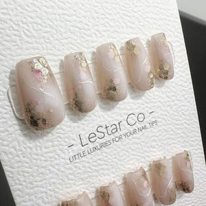 A set of press-on nails featuring a translucent nude base with gold and pink hexagonal glitter accents, white swirl line details, and a glossy finish. The nails are displayed on a white card labeled L