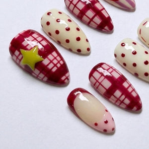 A set of almond-shaped press-on nails featuring a variety of designs including a red and white plaid pattern, cream-colored nails with red polka dots, and a single nail with a yellow star accent over a red plaid background.