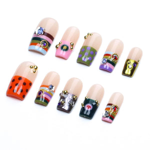 A set of ten press-on nails featuring a nude base with various decorative tips. Designs include horizontal stripes, polka dots, and solid metallic finishes, accented with small gold beads and colorful star-shaped rhinestones.