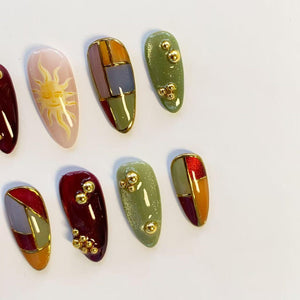 A set of almond-shaped press-on nails featuring various designs including a yellow sun illustration on a pale pink base, geometric color-blocked patterns in red, olive, and grey, and solid olive or deep red nails accented with gold beads.