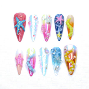 A set of ten stiletto-shaped press-on nails featuring ocean-themed designs. The nails display vibrant gradients, iridescent finishes, and 3D embellishments including miniature starfish, swirling tentacles, and small pearl-like beads.