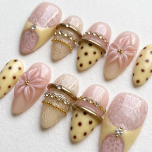 A set of ten almond-shaped press-on nails featuring a palette of pale pink and cream. Designs include intricate lace patterns, raised 3D floral motifs, brown polka dots, and metallic gold rings accented with small clear rhinestones.
