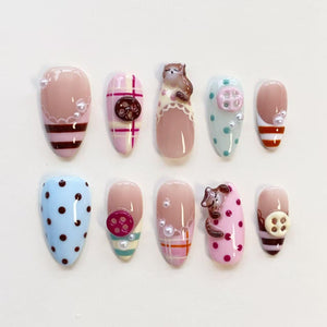 A set of ten press-on nails featuring various designs including pink and brown stripes, plaid patterns, polka dots, small white pearls, and three-dimensional sculpted figures of a cat and a dog, along with attached round buttons.
