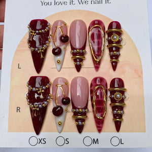 A set of ten press-on nails in a stiletto shape, featuring deep burgundy and nude tones. The designs include gold-toned charms, small white pearls, cherry motifs, and metallic gold accents. The display card includes the text You love it. We nail it.