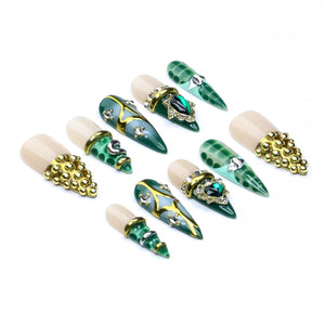A set of ten press-on nails featuring a pointed stiletto shape. The designs include a mix of nude bases, deep green patterns, gold metallic accents, embedded clear rhinestones, and clusters of small gold beads arranged in decorative patterns.