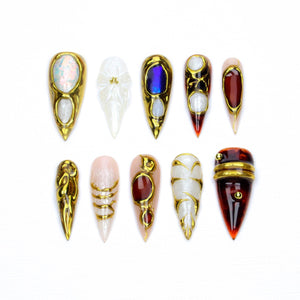 A set of ten stiletto-shaped press-on nails featuring various designs. The nails include gold metallic accents, iridescent finishes, deep red tones, and intricate raised gold detailing on white, clear, and dark brown bases.