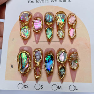 A set of ten press-on nails with a light pink base, each featuring irregular, iridescent abalone shell fragments encased in raised, textured gold-toned metal borders. The display card includes the text L, R, XS, S, M, and L.