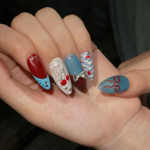 Almond-shaped fingernails featuring intricate nail art designs. The nails display a mix of red, white, and blue colors with patterns including white lace, red cherries, silver bows, glittery stripes, red stars, and a red ribbon motif.