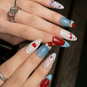 Almond-shaped nails featuring a mix of red, blue, and white lace-patterned polish. The design includes silver bow charms, small red cherry accents, and tiny silver studs. A silver hamsa hand ring is worn on the index finger.