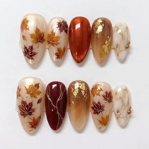 A set of ten almond-shaped press-on nails featuring autumn-themed designs. The nails display cream bases with painted maple leaves in orange and burgundy, solid metallic copper, brown with gold leaf flakes, and white marble patterns.