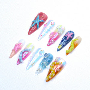 A set of ten stiletto-shaped press-on nails featuring ocean-themed 3D decorations. The designs include colorful starfish, octopus tentacles, pearls, and iridescent finishes in shades of pink, blue, yellow, and white with glossy textures.
