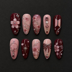 A set of ten almond-shaped press-on nails featuring a color palette of deep burgundy and soft blush pink. The designs include 3D cherry blossom motifs, silver metallic filigree, sparkling rhinestones, a bow charm, and a logo emblem.