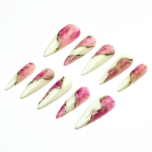A set of ten stiletto-shaped press-on nails featuring a split design. One half is a solid off-white, while the other half displays a pink and white marbled pattern. A thin, metallic gold line separates the two distinct sections.