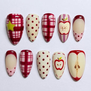 A set of ten press-on nails featuring various designs including red and white plaid patterns, cream backgrounds with red polka dots, a yellow star accent, vertical purple stripes with apple illustrations, and a cross-section of an apple.