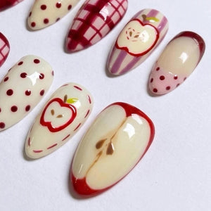 A set of press-on nails featuring various designs including cream-colored nails with dark red polka dots, red plaid patterns, striped backgrounds with red apple illustrations, and a detailed cross-section of a red apple.