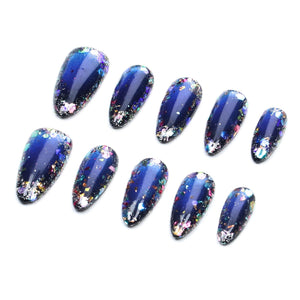 A set of ten almond-shaped press-on nails featuring a deep navy blue base color that transitions into a lighter blue center. The surface is decorated with multi-colored metallic foil flakes in shades of silver, purple, and gold.