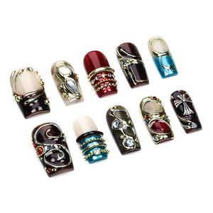 A set of ten press-on nails featuring various designs including deep red, blue, and neutral tones. The nails are decorated with metallic gold accents, embedded rhinestones, and a silver cross emblem on one of the dark brown pieces.