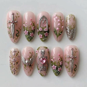 A set of ten almond-shaped press-on nails with a pale pink base. Designs include silver sword charms, pink floral accents, green moss textures, a silver gothic arch, lily of the valley, silver crosses, a winged skull, and a skeleton with a pink heart
