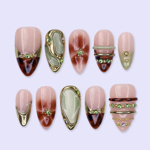 Ten almond press-on nails laid in two rows on a lilac background. Glossy sheer nude bases with brown tortoiseshell swirls, sage green jade-look panels rimmed in raised gold, gold chrome tips, and rows of tiny green gems.