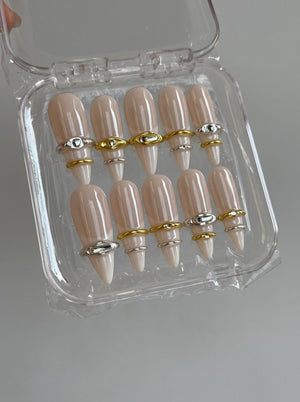 A set of ten almond-shaped press-on nails in a glossy nude beige finish, each adorned with a single metallic ring accent in either gold or silver tones, presented in a clear plastic storage container with a hinged lid.