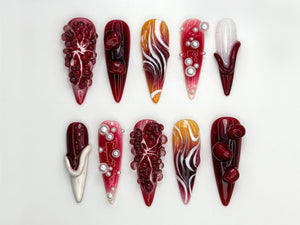 A collection of ten long, pointed artificial nails with various designs. The nails feature a range of colors, including deep reds, oranges, and whites, with intricate patterns and embellishments like