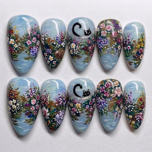 A set of ten almond-shaped press-on nails featuring a hand-painted garden scene with a light blue background. The design includes colorful floral clusters in pink, purple, and white, with a small black cat peeking out on two nails.