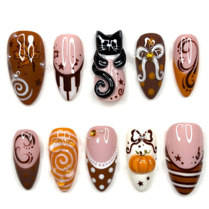 A set of ten press-on nails featuring autumn-themed designs including a black cat, a pumpkin, maple leaves, swirls, polka dots, and musical notes in a color palette of brown, orange, white, and pink with glossy finishes and 3D accents.