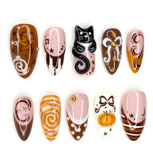 A set of ten press-on nails featuring autumn-themed designs including a brown maple leaf, a black cat figure, a small orange pumpkin, white bows, musical notes, stars, and swirling patterns in shades of brown, orange, white, and pink.