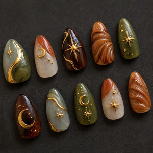A set of ten almond-shaped press-on nails featuring a celestial theme. The designs include deep olive green, brown, and cream marbled patterns accented with gold-toned metal charms shaped like crescent moons and four-pointed stars.