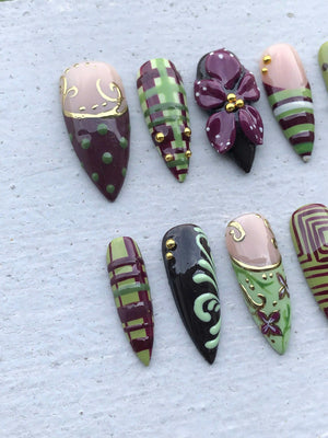 A set of almond-shaped press-on nails featuring a variety of designs including green and burgundy plaid patterns, dark purple floral accents with gold beads, light pink bases with gold swirls, and dark brown surfaces with light green scrolls.