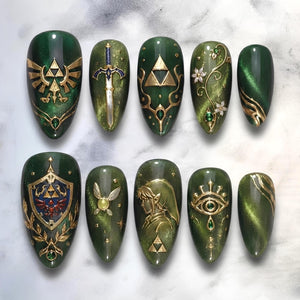 A set of ten almond-shaped press-on nails featuring a deep forest green cat-eye magnetic polish base. The designs include gold-toned emblems, a sword, a shield, a fairy, a profile silhouette, and floral accents with small gems.