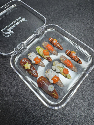 A set of ten press-on nails in a clear plastic case labeled Pinnacle. The nails feature autumn-themed designs including orange pumpkins, white ghosts, brown stripes, maple leaves, and gold accents, with various 3D charms attached.