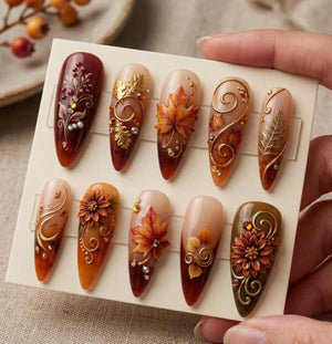 A set of ten almond-shaped press-on nails featuring autumn-themed designs. The nails display warm colors like deep red, orange, and brown, decorated with 3D gold leaf accents, floral motifs, swirling lines, and small embedded crystals.