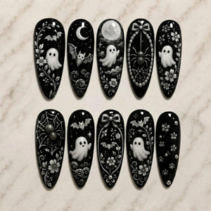 A set of ten almond-shaped press-on nails with a glossy black base. The designs feature white hand-painted motifs including ghosts, bats, spiders, spiderwebs, crescent moons, full moons, bows, floral vines, and small circular accents.