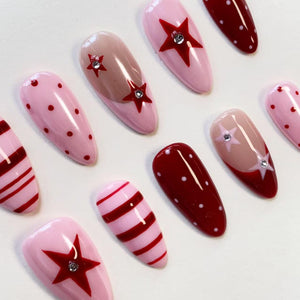 A set of almond-shaped press-on nails featuring a variety of designs including light pink with red polka dots, red with white polka dots, pink and red stripes, and nude bases accented with red stars and small embedded clear rhinestones.