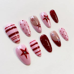 A set of ten almond-shaped press-on nails featuring a variety of designs in shades of red and light pink. The patterns include horizontal stripes, polka dots, and red star motifs accented with small, clear, circular rhinestones.