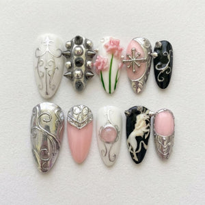 A set of ten press-on nails featuring diverse designs including silver filigree on white, metallic studs, pink floral accents, a silver cross, black with silver swirls, a solid silver finish, pink with silver armor, and a unicorn graphic.