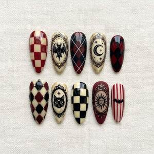 A set of ten almond-shaped press-on nails featuring various designs including red and cream checkerboard, black and red argyle, vertical stripes, and ornate oval frames containing illustrations of a bat, moon, cat, and sun.