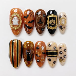 A set of ten almond-shaped press-on nails featuring Halloween and autumn themes. Designs include a white ghost in a gold frame, black bats, spider webs, a witch hat, a jack-o'-lantern, a black cat, soot sprites, and dried flowers.