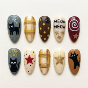A set of ten almond-shaped press-on nails featuring various designs including black cats, yellow plaid patterns, brown polka dots, gold stars, a red star, and the text MEOW MEOW and STAR CAT painted in black on cream and gray backgrounds.