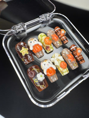 A set of ten press-on nails arranged in a clear plastic case. The designs feature autumn themes including 3D orange pumpkins, white cats, maple leaves, plaid patterns, and small white flowers on brown, cream, and clear base colors.
