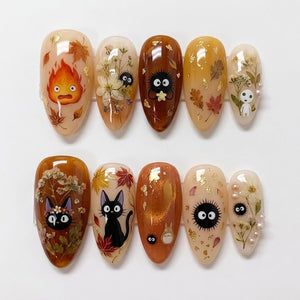 A set of ten almond-shaped press-on nails featuring autumn-themed designs, including illustrations of a fire spirit, black cats, soot sprites, and forest spirits, accented with dried flowers, gold leaf flakes, and small white pearls.