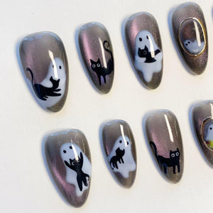 A set of almond-shaped press-on nails featuring a dark grey magnetic cat-eye finish with glitter. The designs include white ghost silhouettes and black cat illustrations, with one nail featuring a small gold-toned oval frame accent.
