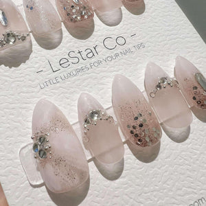 A set of almond-shaped press-on nails in a translucent pale pink finish, featuring silver glitter accents and embedded clear rhinestones. The nails are displayed on a white textured card labeled LeSta